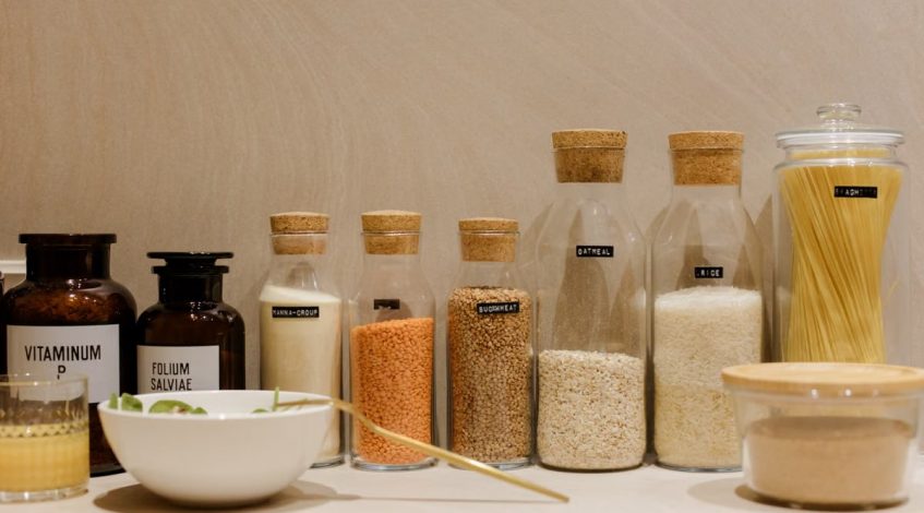 A variety of grains and pasta in labeled glass bottles displayed on a kitchen counter.